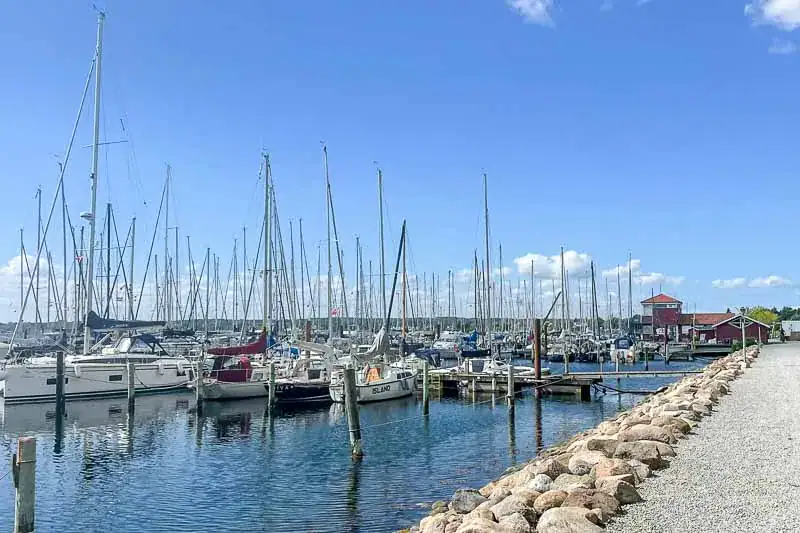 Havn med sejlbåde parkeret langs kajen, blå himmel og lette skyer i baggrunden. Stien er belagt med grus, og rækker af både ses til venstre og højre.