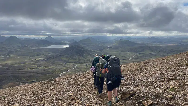 Gruppe vandrere med rygsække på stenrig bjergsti med udsigt over grønne dale, floder og fjerne bjerge under dramatisk skyet himmel