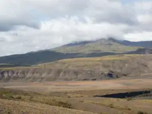 Islandsk landskab med fladt terræn, eroderede klipper og skyklædt bjerg i baggrunden – naturskøn udsigt langs Laugavegur-ruten