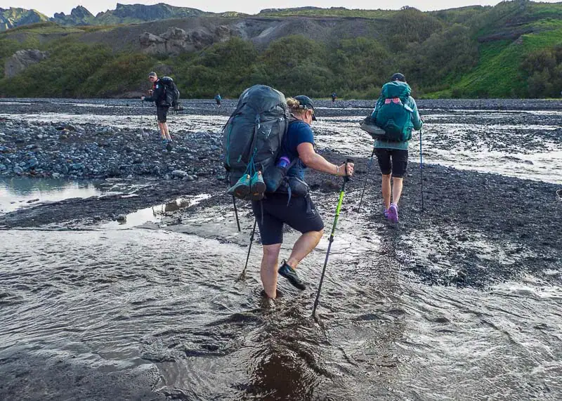 Vandrere krydser lavvandet elv i Island med rygsække og vandrestave – aktiv trekkingoplevelse i rå og naturskøn natur