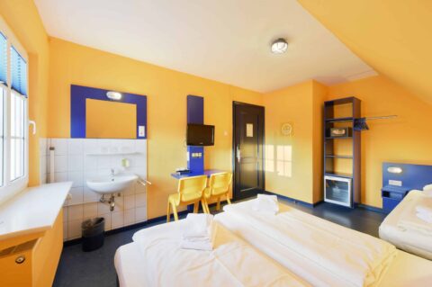 Basic-Zimmer mit drei Betten, einem Schreibtisch, zwei gelben Stühlen, TV und einem Waschbecken. Ein offener Schrank mit Safe und Kühlschrank.