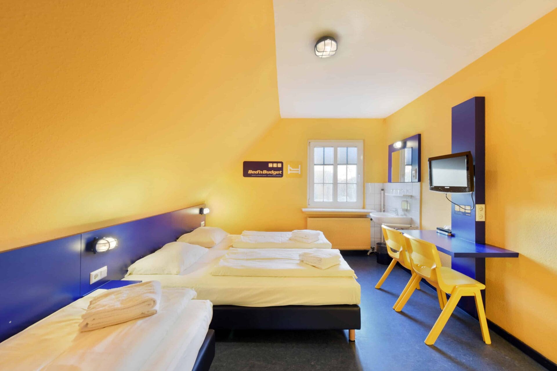 Basic-Zimmer mit drei Betten, einem Schreibtisch, zwei gelben Stühlen, TV, Waschbecken, Fenster und Dachschräge.
