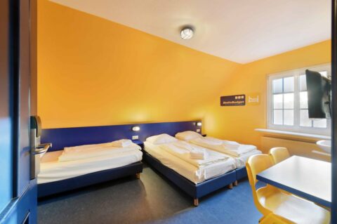 Basic-Zimmer mit drei Betten, einem Schreibtisch, zwei gelben Stühlen, TV, Waschbecken, Fenster und Dachschräge.