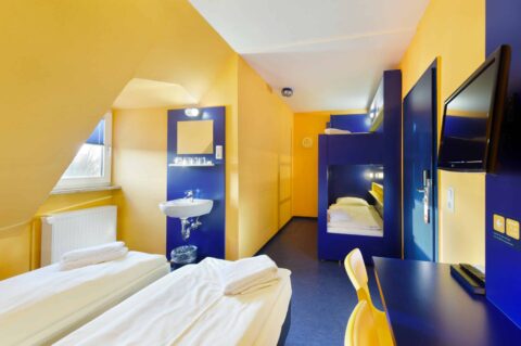 Basic-Zimmer mit einem Etagenbett und zwei Einzelbetten für 4 Personen, einem Schreibtisch, TV, Fenster und einem Waschbecken.