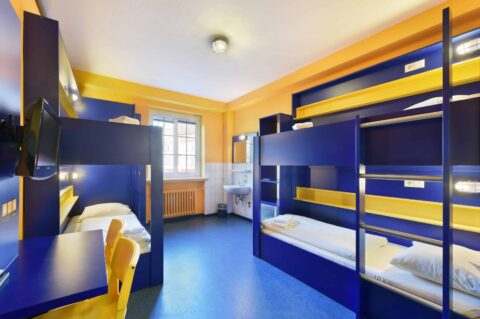 Basic-Zimmer mit zwei Etagenbetten für 4 Personen, einem Schreibtisch, zwei gelben Stühlen, TV, Fenster und einem Waschbecken.