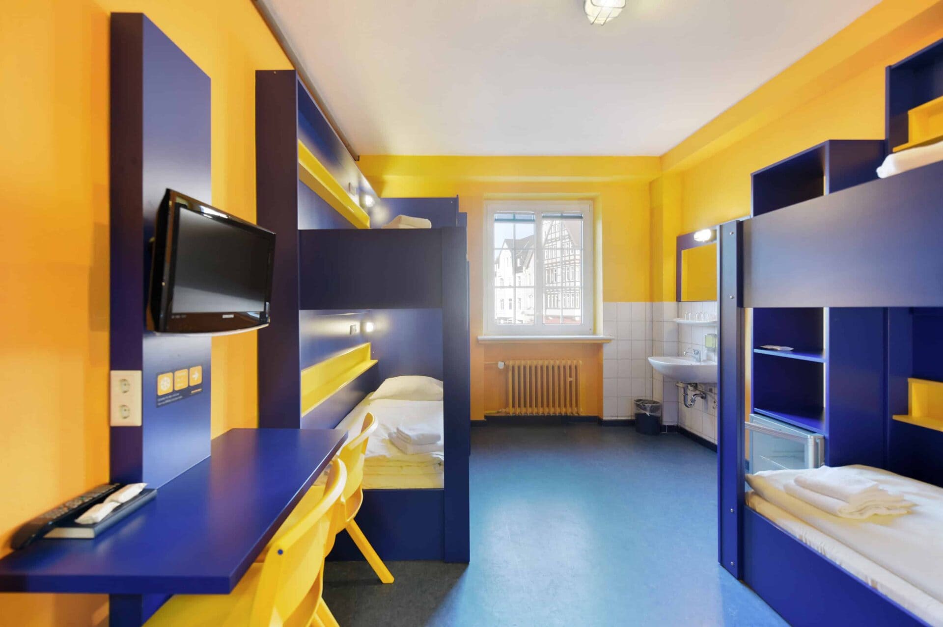 Basic-Zimmer mit zwei Etagenbetten für 4 Personen, einem Schreibtisch, zwei gelben Stühlen, TV, Fenster und einem Waschbecken.