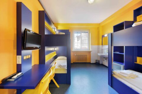 Basic-Zimmer mit zwei Etagenbetten für 4 Personen, einem Schreibtisch, zwei gelben Stühlen, TV, Fenster und einem Waschbecken.
