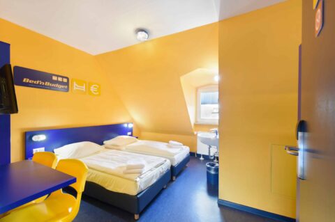 Basic-Zimmer mit einem Doppelbett, einem Einzelbett, einem Schreibtisch, zwei gelben Stühlen, Fenster, Waschbecken und Dachschräge neben dem Bett.