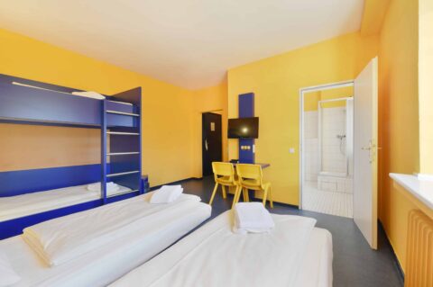 Comfort-Zimmer mit zwei Einzelbetten und einem Etagenbett für 4 Personen, Schreibtisch, TV, zwei Stühle und eigenem Badezimmer.
