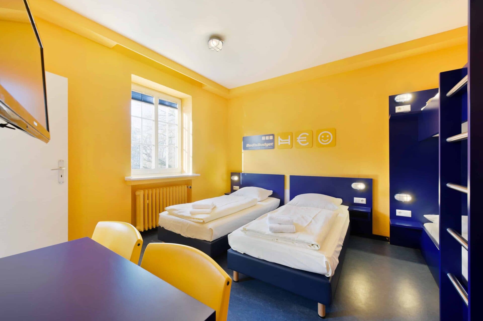 Comfort-Zimmer mit zwei Einzelbetten und einem Etagenbett für 4 Personen, Fenster, TV, Schreibtisch und zwei Stühlen.