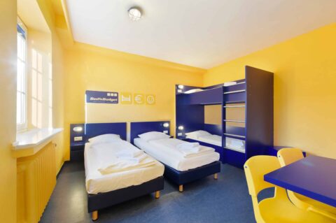 Comfort-Zimmer mit zwei Einzelbetten und einem Etagenbett für 4 Personen, Fenster und Schreibtisch mit Stuhl.