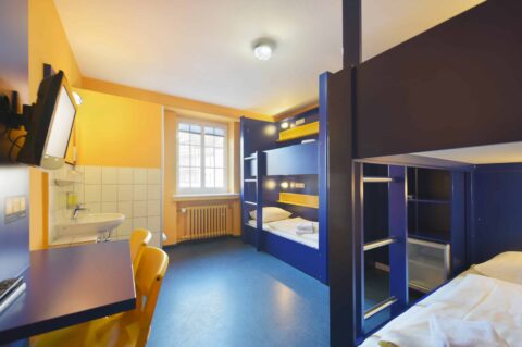 Economy-Zimmer mit zwei Etagenbetten für 4 Personen, einem Schreibtisch, zwei Stühlen, TV, Fenster und einem Waschbecken.