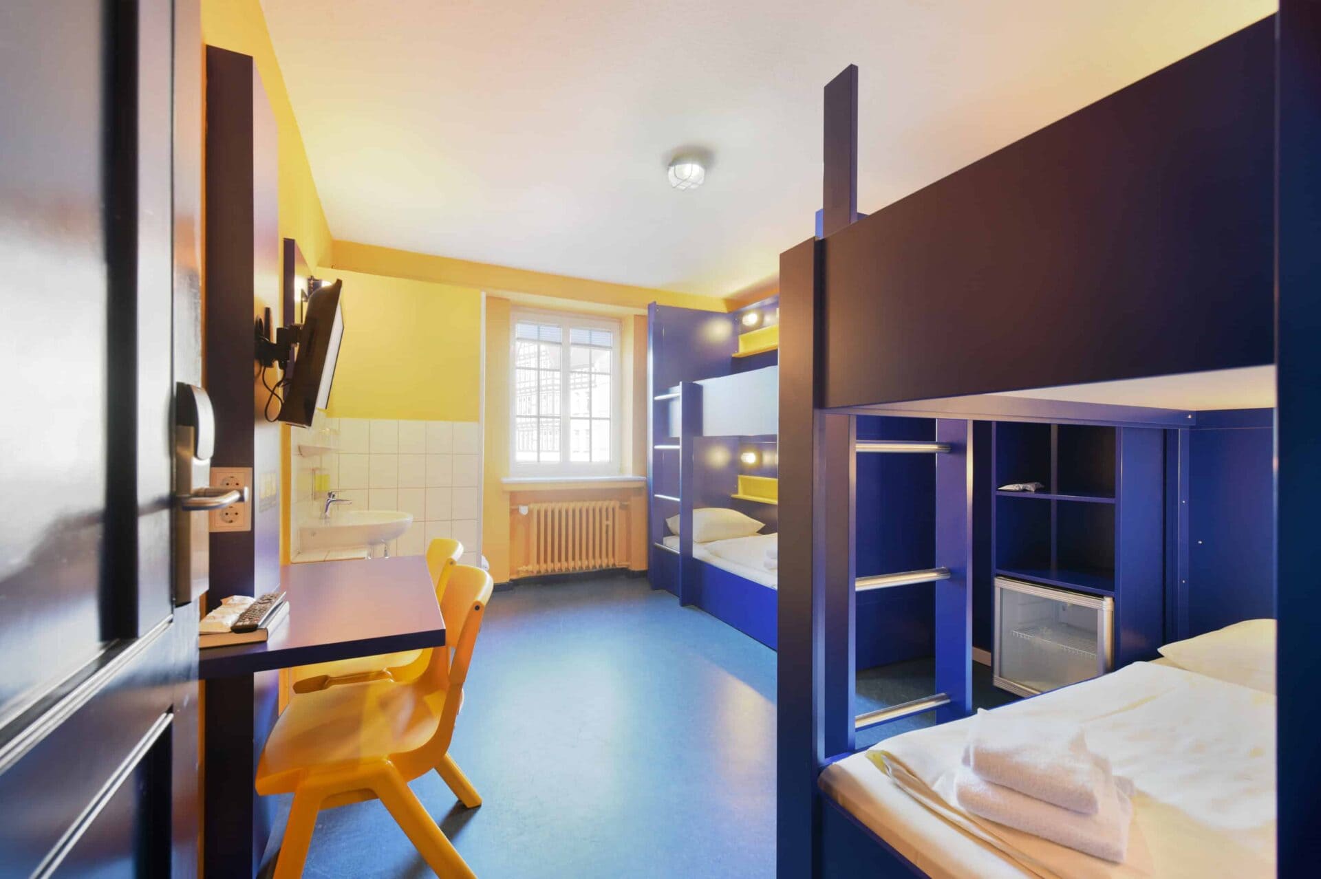 Economy-Zimmer mit zwei Etagenbetten für 4 Personen, einem Schreibtisch, zwei Stühlen, TV, Fenster und einem Waschbecken.