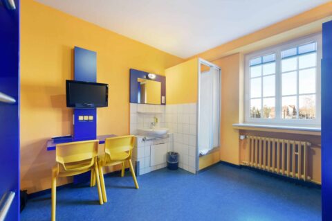Economy Zimmer mit Schreibtisch, zwei Stühlen, TV, Fenster, Waschbecken und eigener Dusche.
