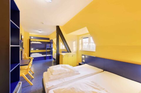 Basic-Zimmer mit einem Etagenbett, einem Doppelbett und einem Einzelbetten für 5 Personen. Ein Schreibtisch, Stühle, Schrank und Dachschräge mit Fenster.