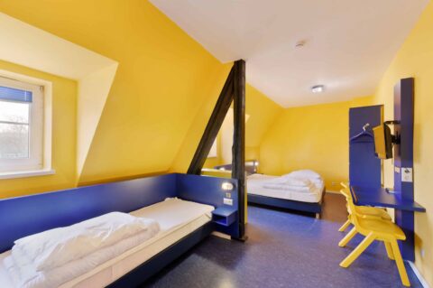 Basic-Zimmer mit drei Einzelbetten, einem Schreibtisch, zwei Stühle, TV und Dachschräge mit Fenster.