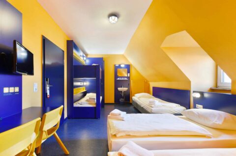 Basic-Zimmer mit einem Etagenbett und zwei Einzelbetten, einem Schreibtisch, zwei Stühle, TV, Fenster, Waschbecken und Dachschräge.