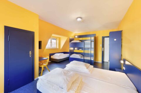 Basic-Zimmer mit einem Etagenbett und drei Einzelbetten für 5 Personen, einem Schreibtisch, Stuhl, Schrank, Fenster.