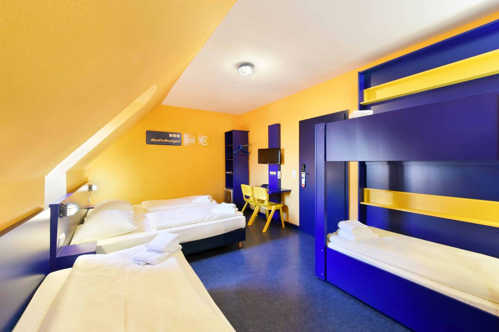 Basic-Zimmer mit einem Etagenbett und drei Einzelbetten für 5 Personen, einem Schreibtisch, zwei Stühle, Schrank, TV und Dachschräge.
