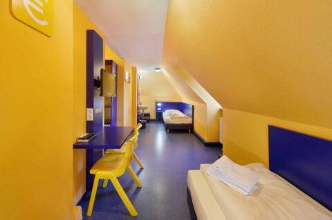 Basic-Doppelzimmer mit zwei Einzelbetten, einem Schreibtisch, zwei Stühlen, TV, Waschbecken und Dachschräge.