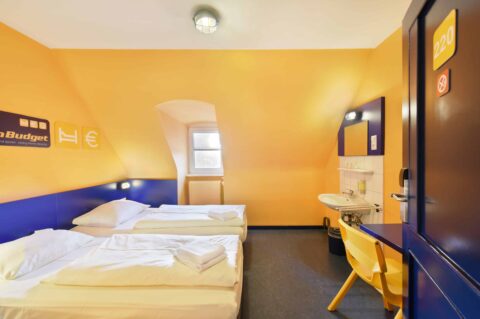 Basic-Doppelzimmer mit zwei Einzelbetten, einem Stuhl, Fenster, Waschbecken und Dachschräge.
