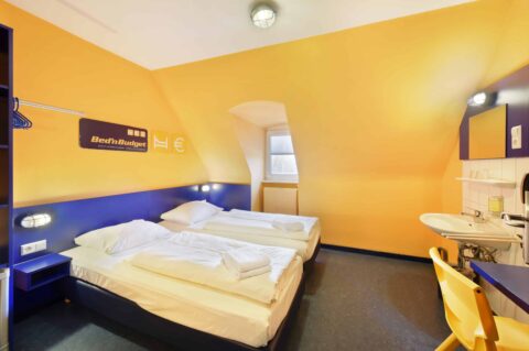 Basic-Doppelzimmer mit zwei Einzelbetten, einem Stuhl, Fenster, Waschbecken und Dachschräge.