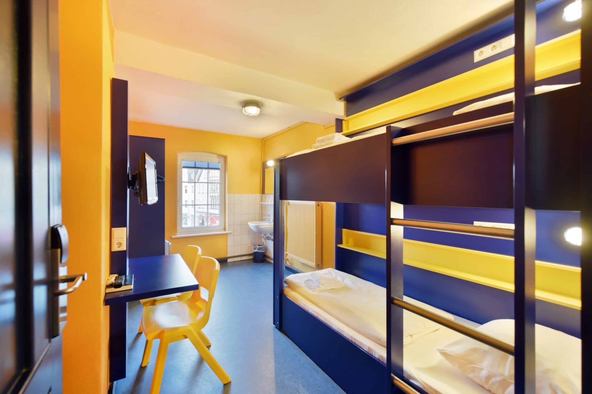 Basic-Doppelzimmer mit einem Etagenbett für zwei Personen , einem Schreibtisch, zwei Stühlen, TV und Waschbecken.
