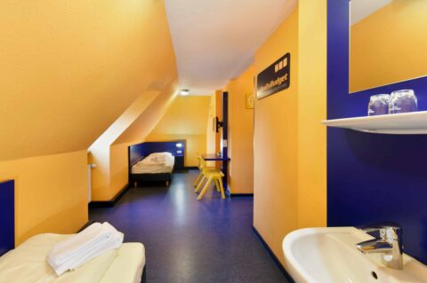 Basic-Doppelzimmer mit zwei Einzelbetten, einem Schreibtisch, zwei Stühlen, TV, Waschbecken und Dachschräge.