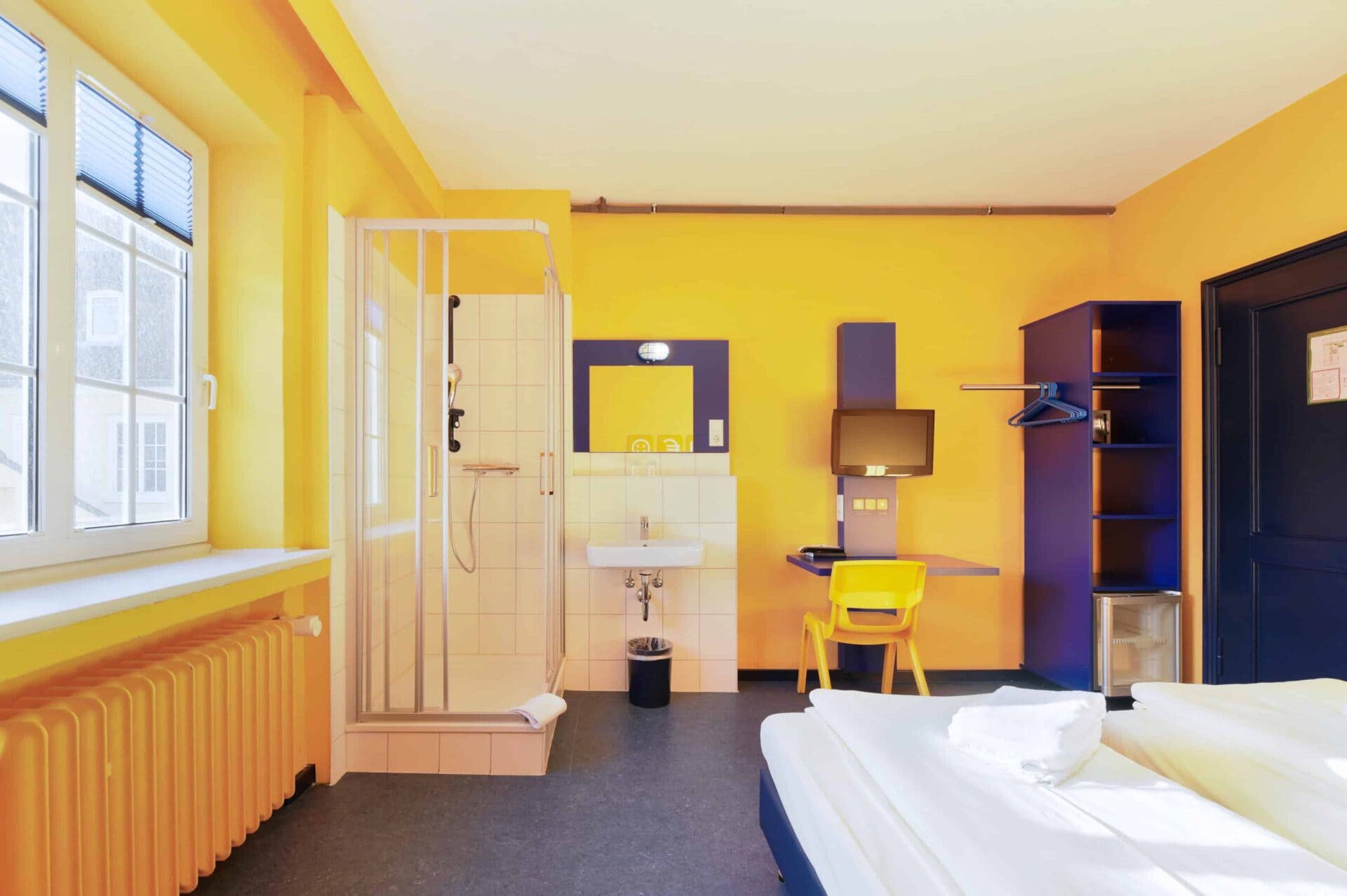 Economy-Zimmer mit zwei Einzelbetten, einem Schreibtisch, einem Stuhl, TV, Fenster, Schrank mit Kühlschrank, Waschbecken und eigener Dusche.