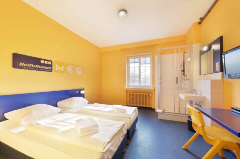 Economy-Zimmer mit zwei Einzelbetten, einem Schreibtisch, einem Stuhl, TV, Fenster, Waschbecken und eigener Dusche.