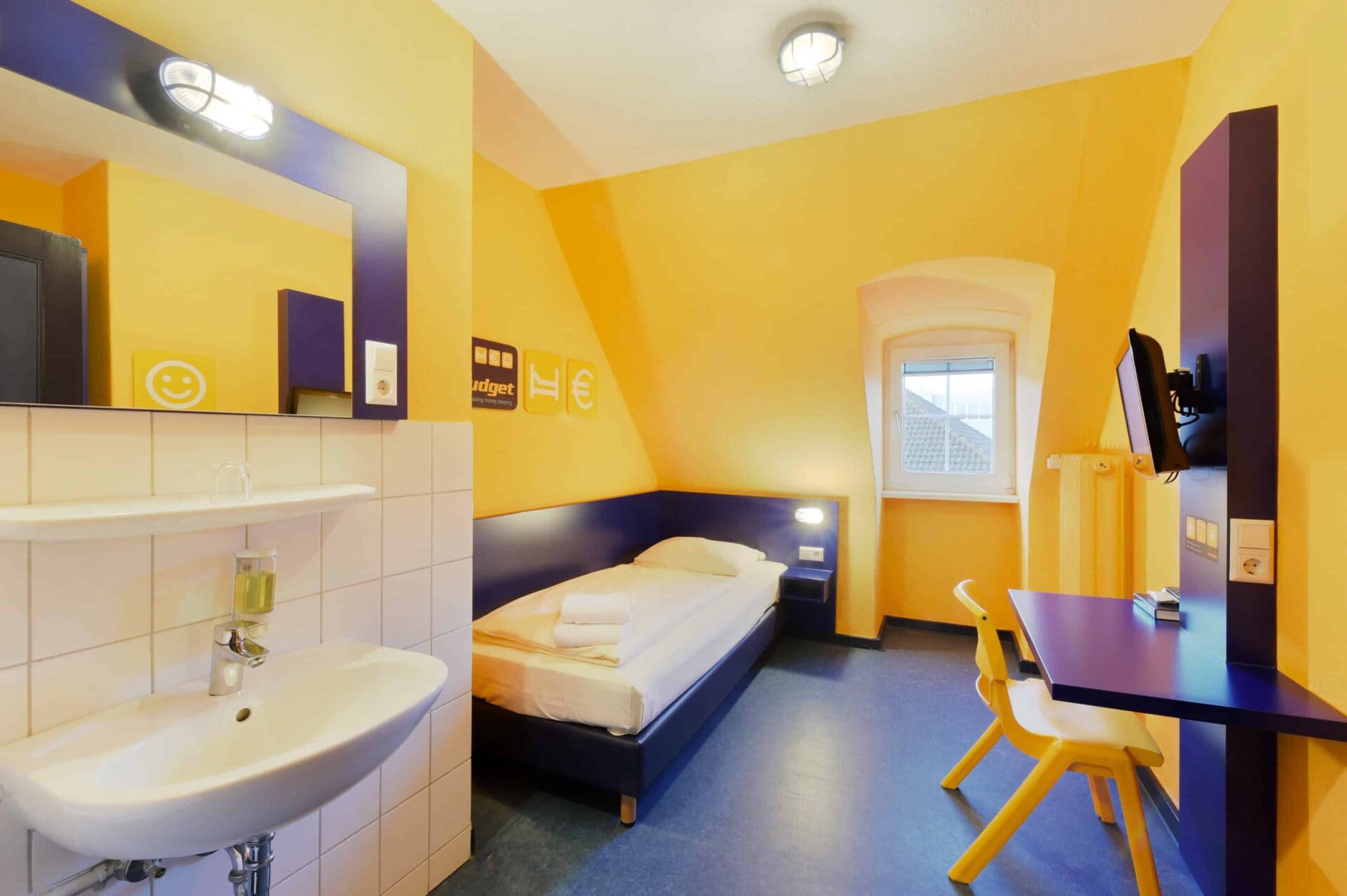 Basic-Einzelzimmer mit einem Einzelbett, einem Schreibtisch, einem Stuhl, TV, Fenster, Waschbecken und Dachschräge.
