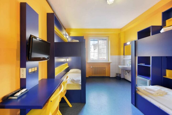 Zimmer mit zwei Etagenbetten für 4 Personen, einem Schreibtisch, zwei gelben Stühlen, TV, Fenster und einem Waschbecken.