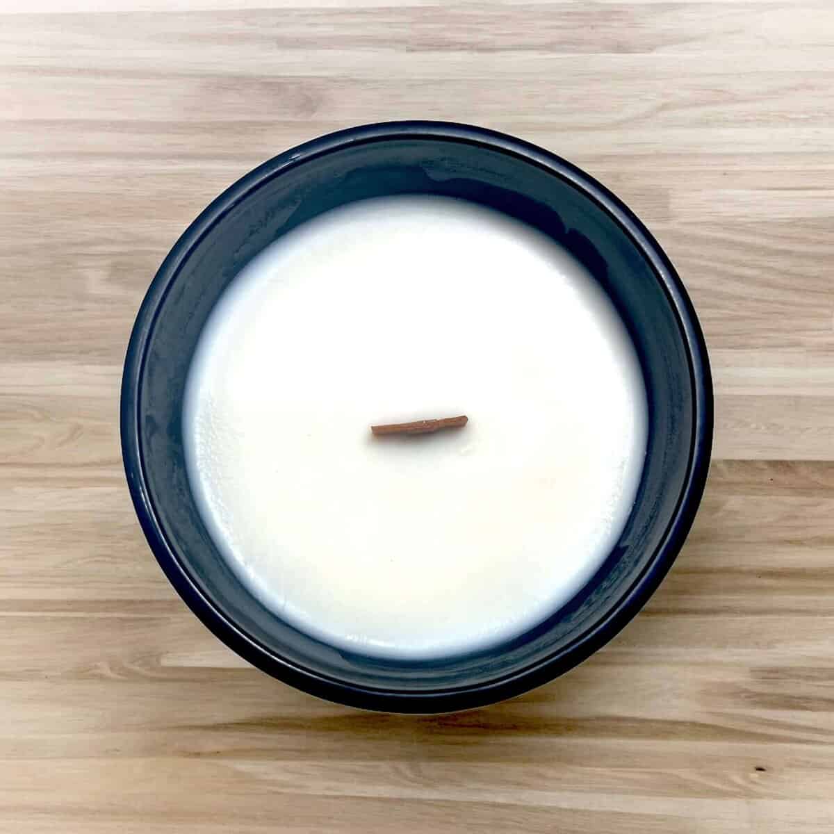 Close-up of a white soy wax candle in a black jar, with a wooden wick, on a wooden surface.