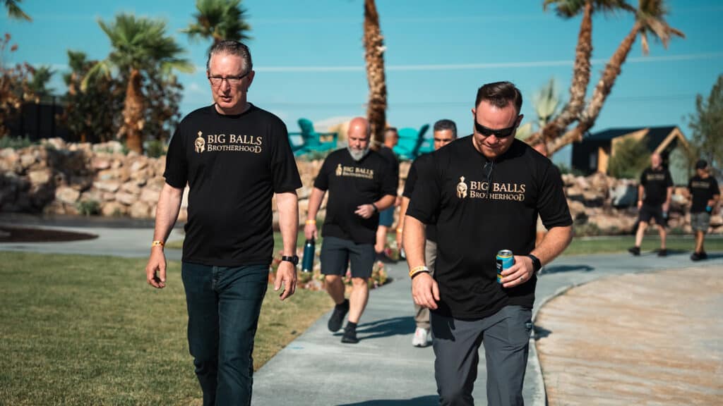 Big Balls Brotherhood men walking outdoors, wearing black T-shirts with the logo, at a social gathering in a sunny, scenic location.