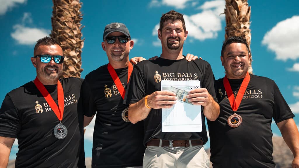 Medal-winning members of the Big Balls Brotherhood team celebrating outdoors after competition, wearing matching black shirts with a Spartan helmet logo, under bright blue skies.