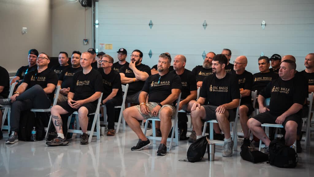 Men attending a Big Balls Brotherhood event, seated in a conference room, wearing matching black T-shirts with the organization’s logo, engaged in a gathering or seminar.