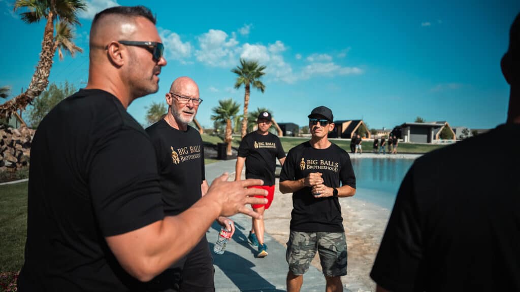 Bearded men wearing black "Big Balls Brotherhood" t-shirts gather outdoors by a pool under a clear blue sky, engaging in conversation at a social event in a sunny, modern residential area.