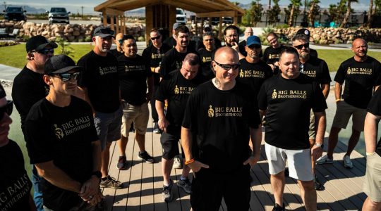 Big Balls Brotherhood members gathered outdoors wearing matching black T-shirts with the group's logo, showcasing camaraderie and brotherhood in a sunny, landscaped setting.