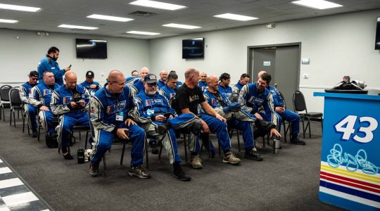 Racing team members sitting in a waiting room, dressed in blue and gray racing suits with NASCAR logos, preparing for a race event, representing the Big Balls Brotherhood.