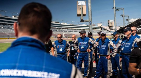 High-energy NASCAR race team members in blue racing suits gathered at a speedway during a race event.