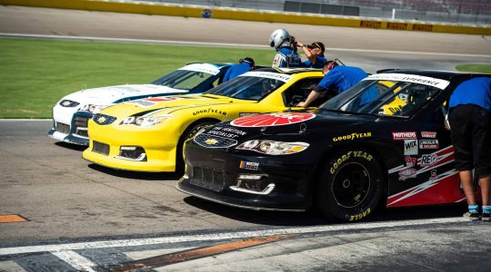 NASCAR race cars lined up at the starting grid with team members preparing for a race at the Big Balls Brotherhood event.