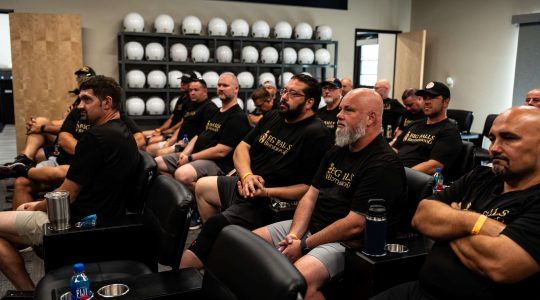 Big Balls Brotherhood members attending a motorcycle club meeting or event, seated in a conference room with motorcycle helmets in the background, showcasing camaraderie and brotherhood.
