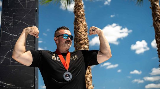 Strong man flexing biceps at Big Balls Brotherhood event with palm trees and blue sky in background.