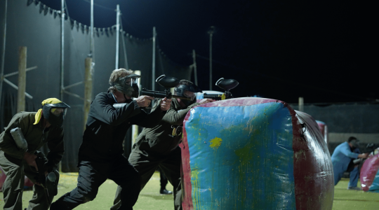 Players engaged in nighttime paintball game, hiding behind colorful inflatable barriers, aiming and shooting paintball guns during a tactical match at Big Balls Brotherhood.