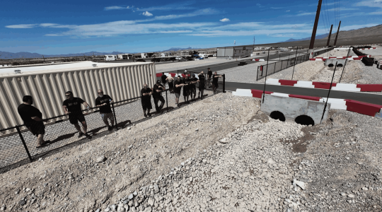 Men in black shirts standing along a chain-link fence at an outdoor go-kart racing track in a desert environment.