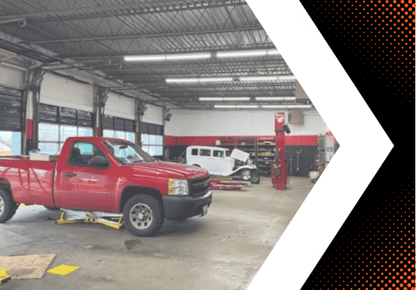 Auto repair shop with red pickup truck on lift and classic cars inside garage.