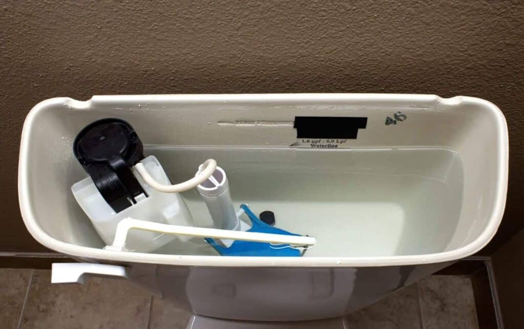 An open toilet tank displays the water level, fill valve, float, and flush valve components in detail as part of Bonfe’s plumbing services for Bonfe Insider, July 2021. The setup is shown against a beige wall backdrop.