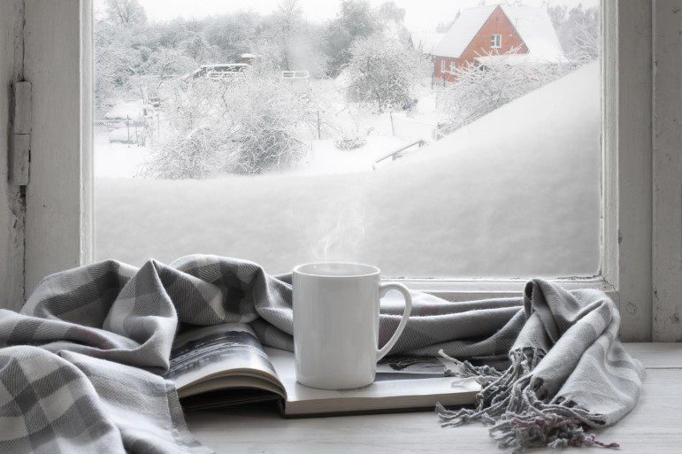 A cozy white mug and an open book rest on a windowsill, draped with a stylish gray plaid blanket. Through the window, freshly fallen snow blankets the ground and trees, creating a warm, welcoming atmosphere—just the kind of inviting scene we’re proud to share in Bonfe Insider, October 2021.