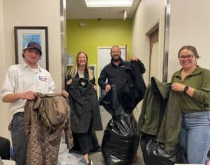 Bonfe team members stand together indoors, smiling as they display coats and jackets collected for the January 2024 Bonfe Insider. Large bags filled with donated clothing are placed at their feet, highlighting their commitment to community service.