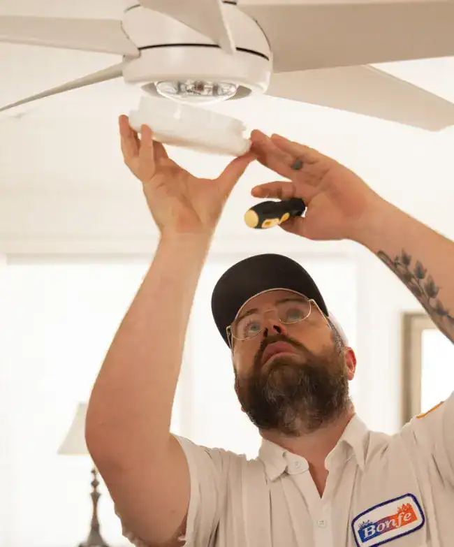 A Bonfe technician, dressed in a company shirt, skillfully repairs or installs a ceiling fan by using a screwdriver and inspecting the fixture above as part of Bonfe’s trusted lighting services.